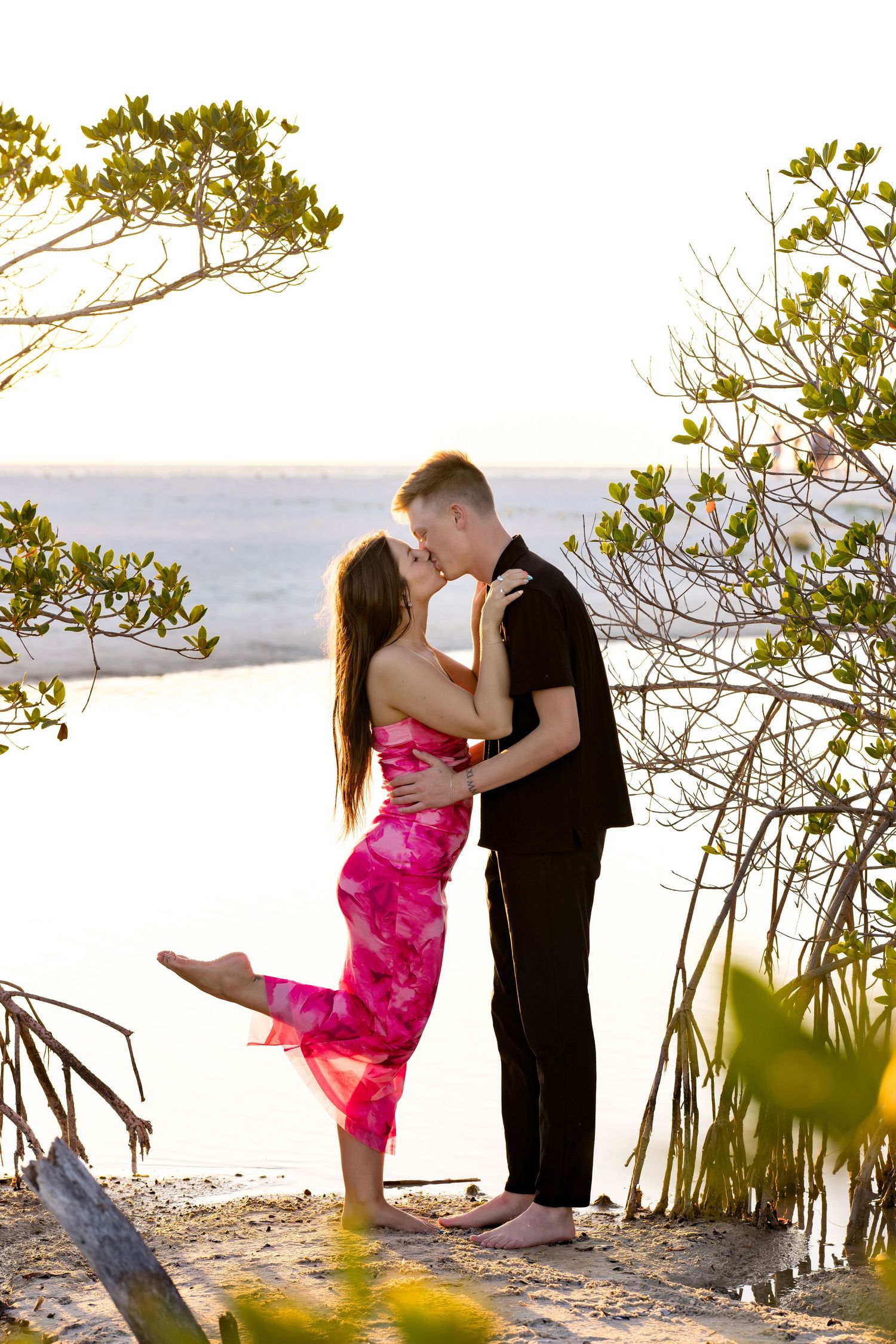 Couple sharing a romantic kiss on a sandy beach at sunset, surrounded by lush green mangrove trees.