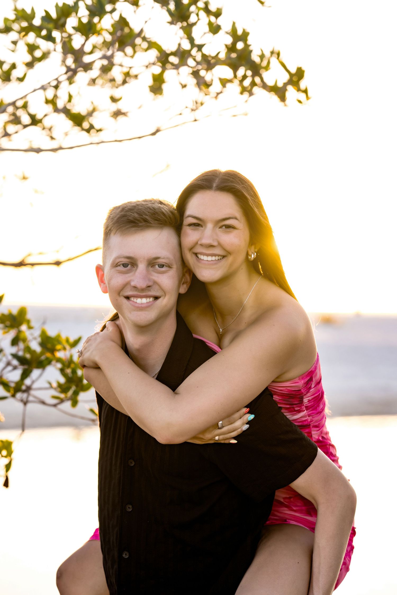 Happy couple sharing a piggyback ride at sunset near the water, woman in pink dress embracing man in black shirt.