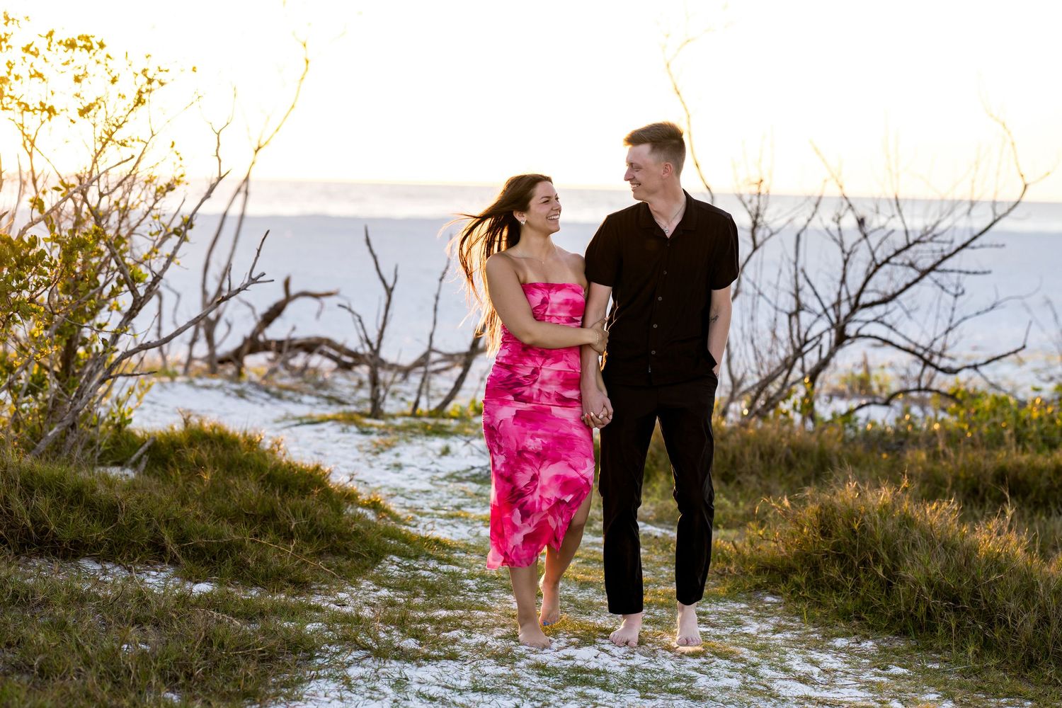 Couple walking barefoot on sandy beach path, woman in pink floral dress, man in black outfit, golden hour lighting.