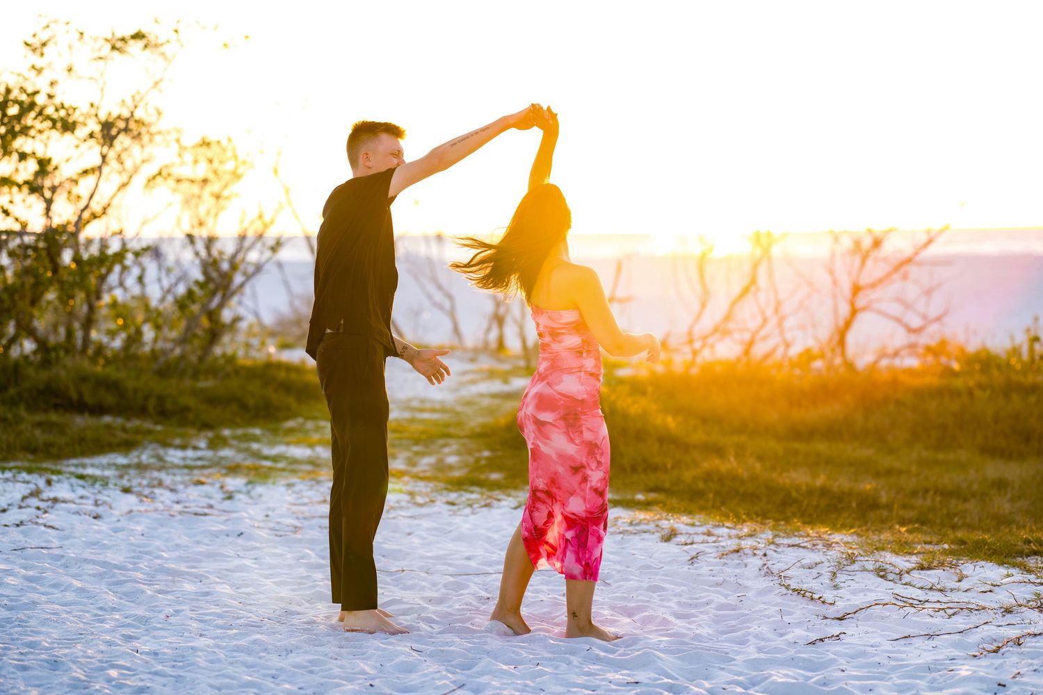 Couple dancing barefoot on sandy beach at golden sunset, man spinning woman in pink floral dress.