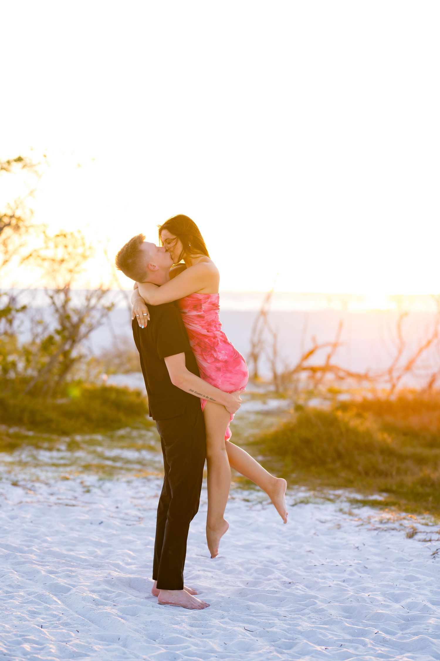 Couple sharing romantic sunset moment on white sand beach, man lifting woman in pink dress with golden backlight.