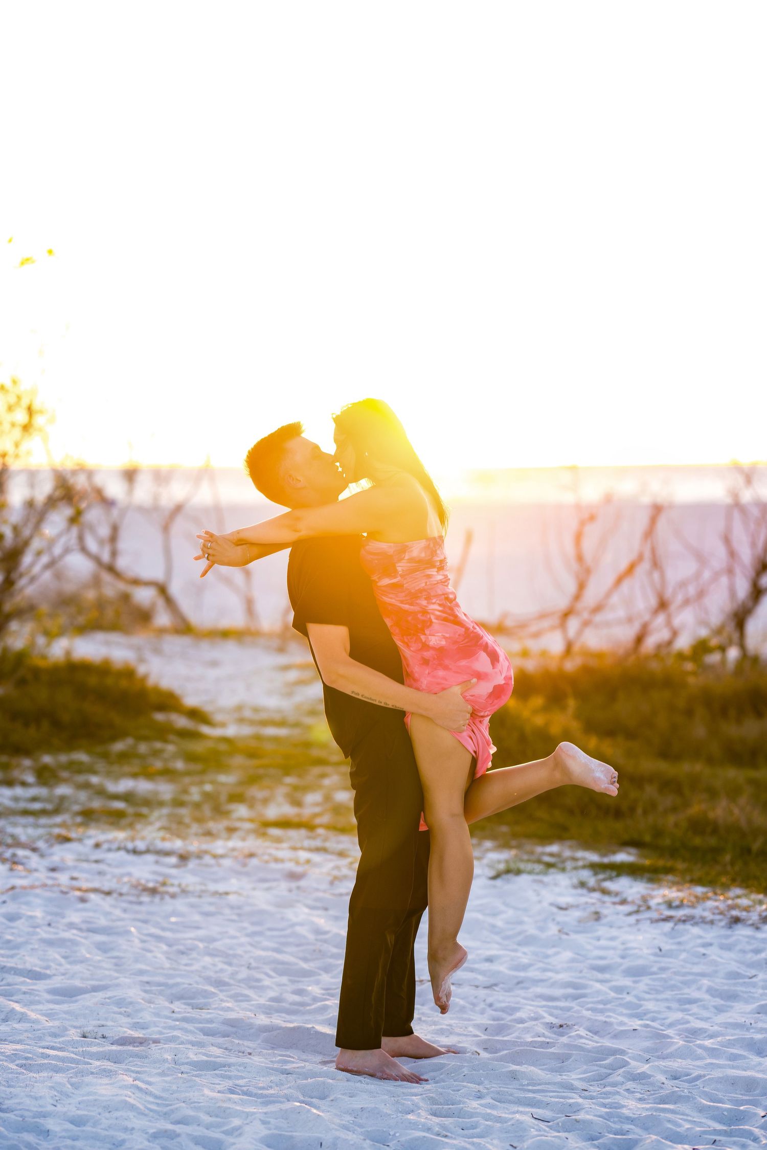 Couple sharing a romantic kiss at sunset on a sandy beach, man lifting woman in pink dress in golden light.