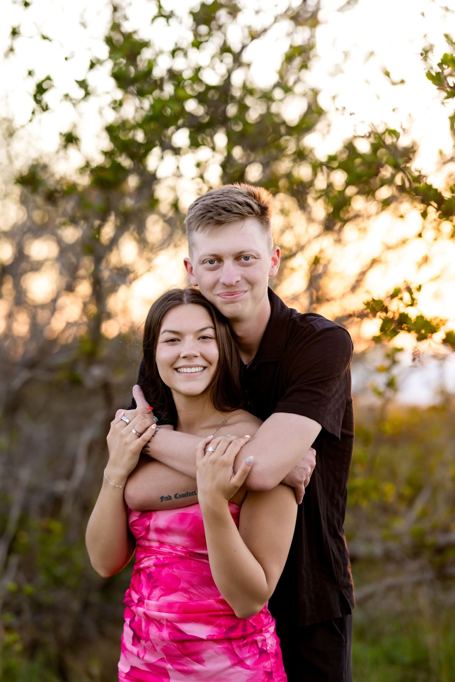 A smiling couple embraces outdoors at golden hour, woman in pink dress, man in black shirt, with trees in background.
