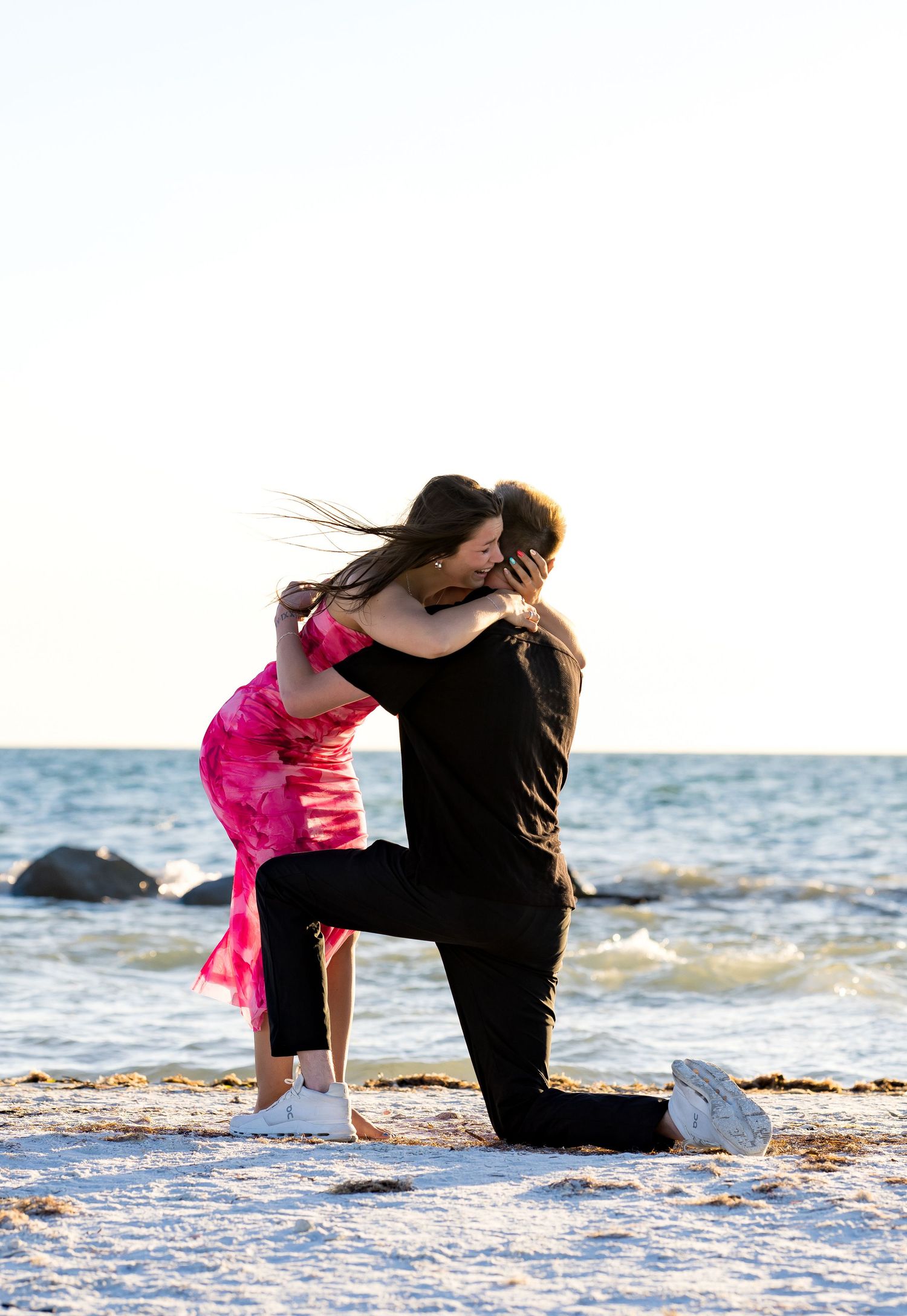Couple sharing romantic beach proposal moment, man kneeling on sandy shore embracing woman in pink dress.