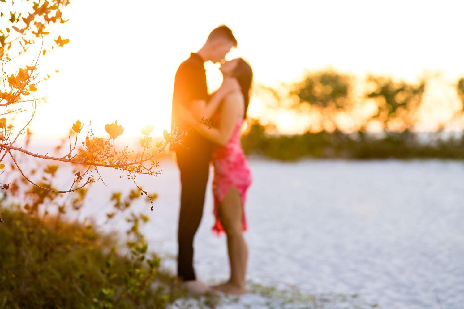 Couple embracing at golden sunset on a beach, backlit by warm sunlight with lush greenery in soft focus.