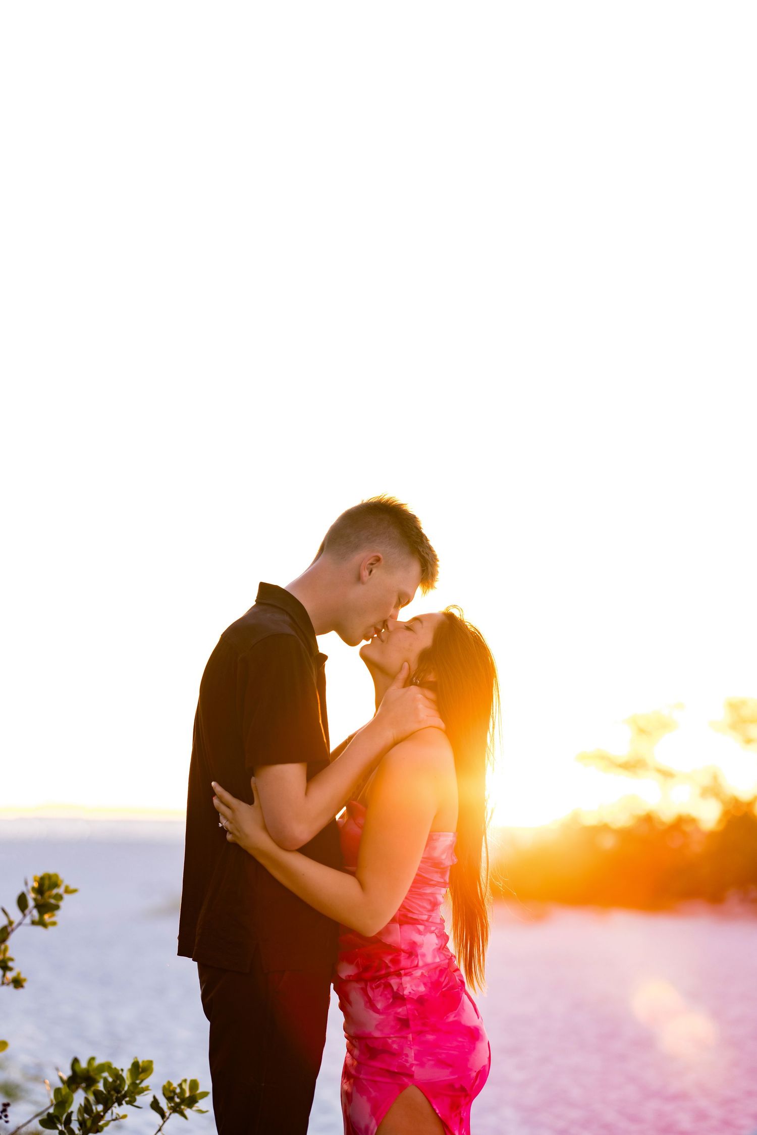 Couple embracing at sunset on a beach, backlit by golden light, man in black shirt and woman in pink dress.