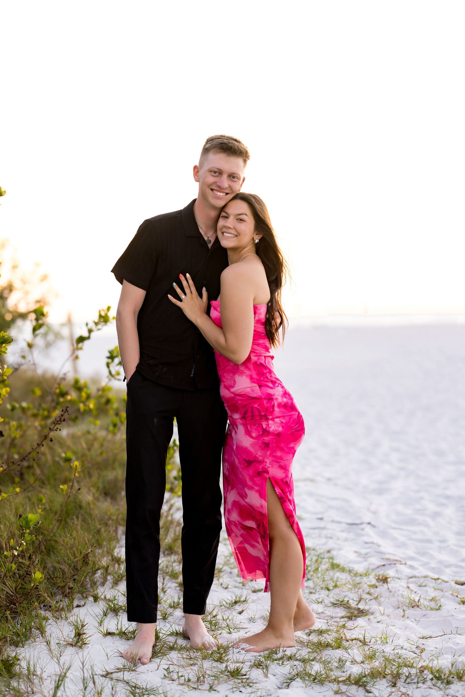 A couple embraces on a white sand beach, woman in pink dress, man in black outfit, smiling at camera during golden hour.