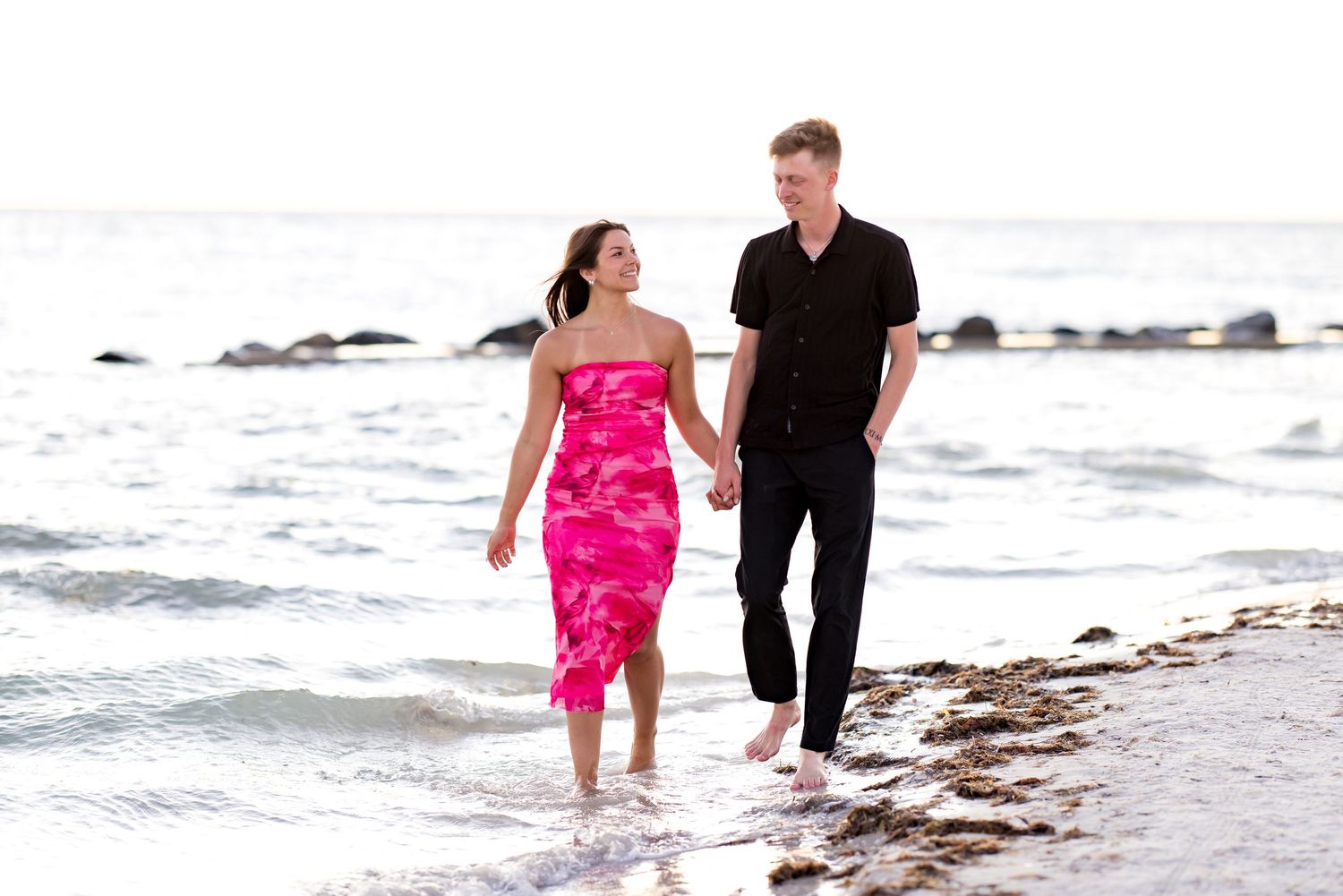 Couple holding hands walking barefoot along a sandy beach shoreline, woman in pink dress, man in black outfit.