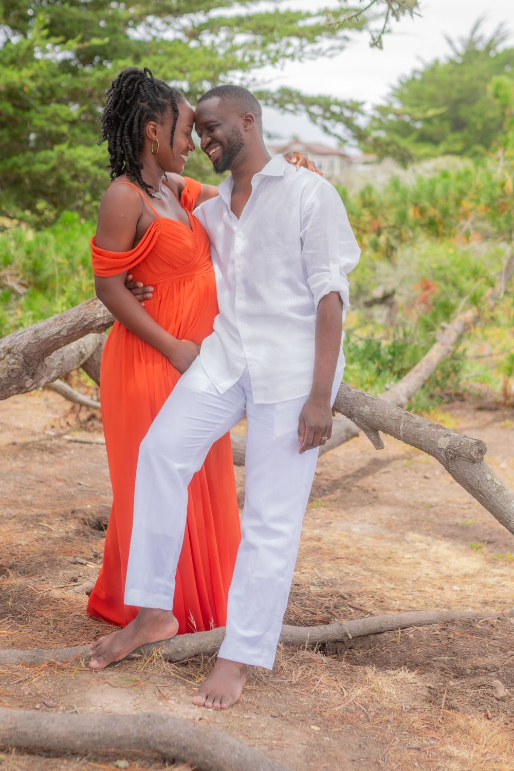 Happy couple posing outdoors, woman in orange maternity dress and man in white outfit, surrounded by trees.