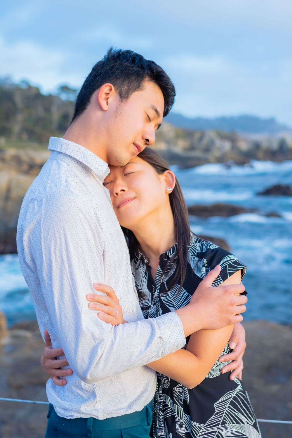 Asian couple sharing tender embrace near rocky ocean coastline at dusk.