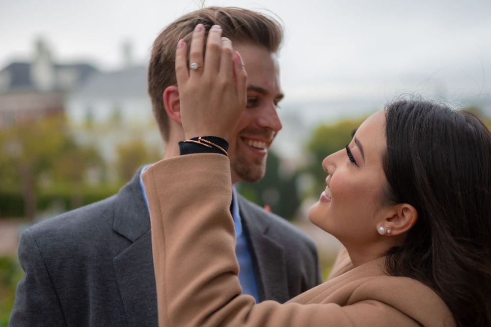 A smiling couple shares a romantic moment outdoors, woman tenderly touching man's face, city skyline in background.