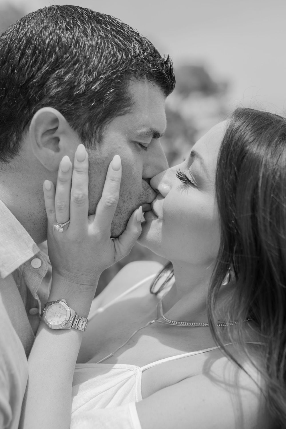 Black and white photo of a couple sharing a romantic kiss, woman holding man's face, wearing engagement ring and watch.