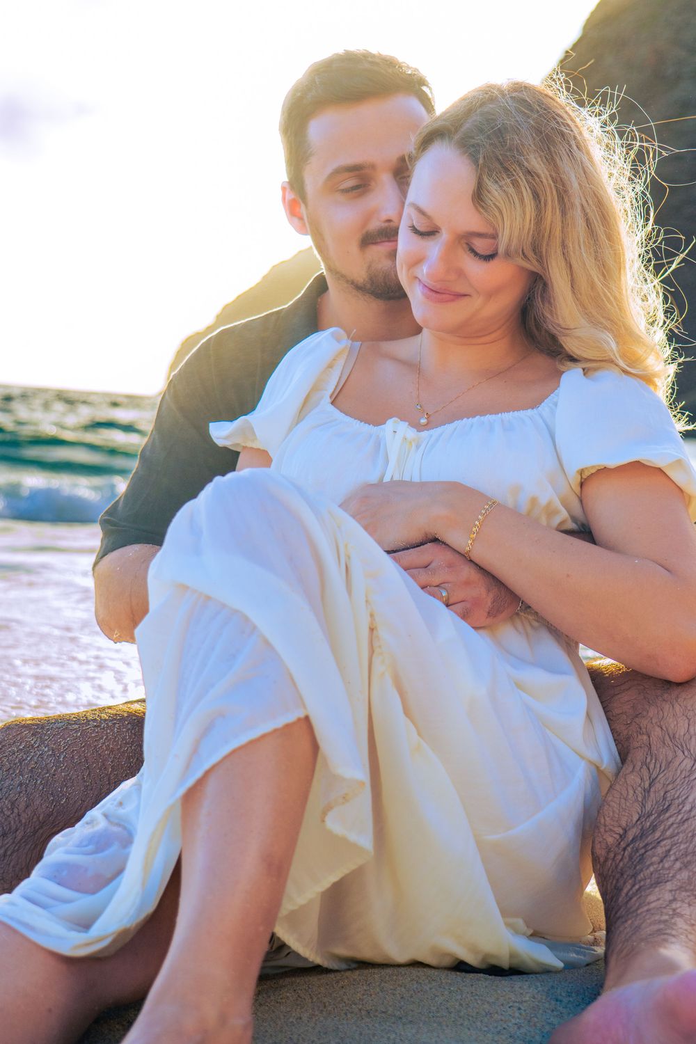 A romantic couple embraces on a beach at sunset, woman in white dress leaning against man in warm golden light.