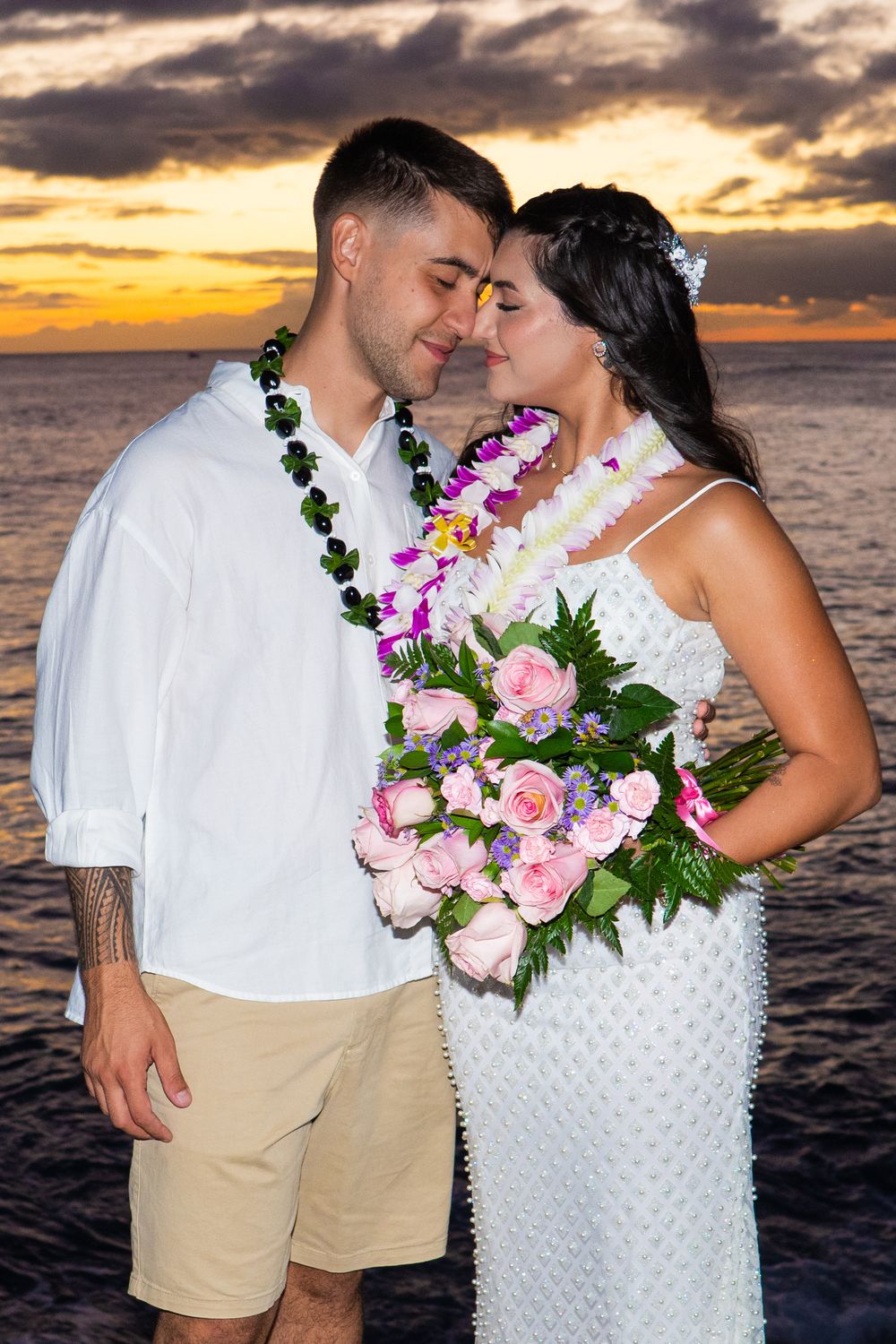 Couple in Hawaiian leis sharing romantic sunset moment at beach wedding, bride holding pink rose bouquet.