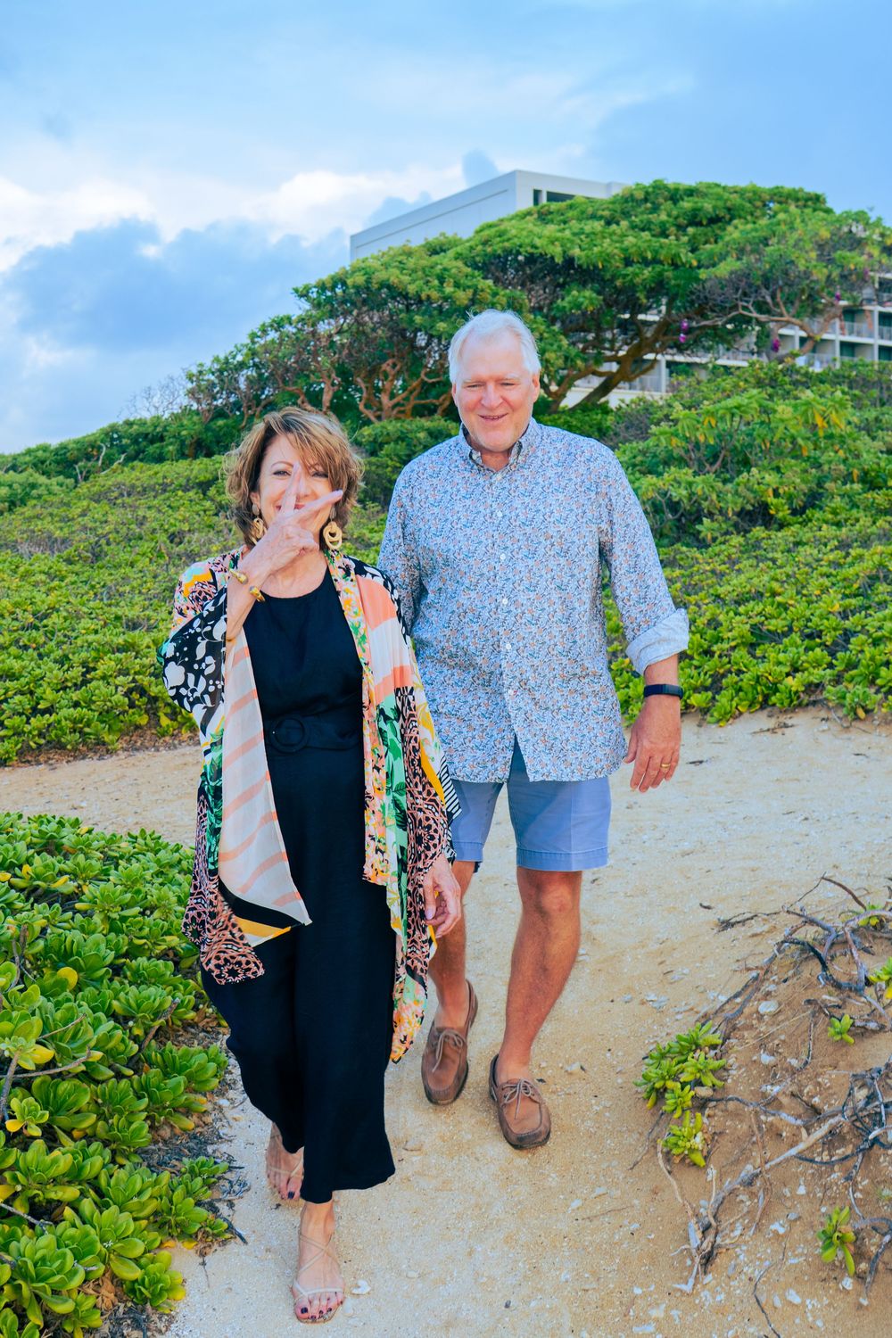 Smiling older couple walking together on a sandy coastal path near lush greenery.