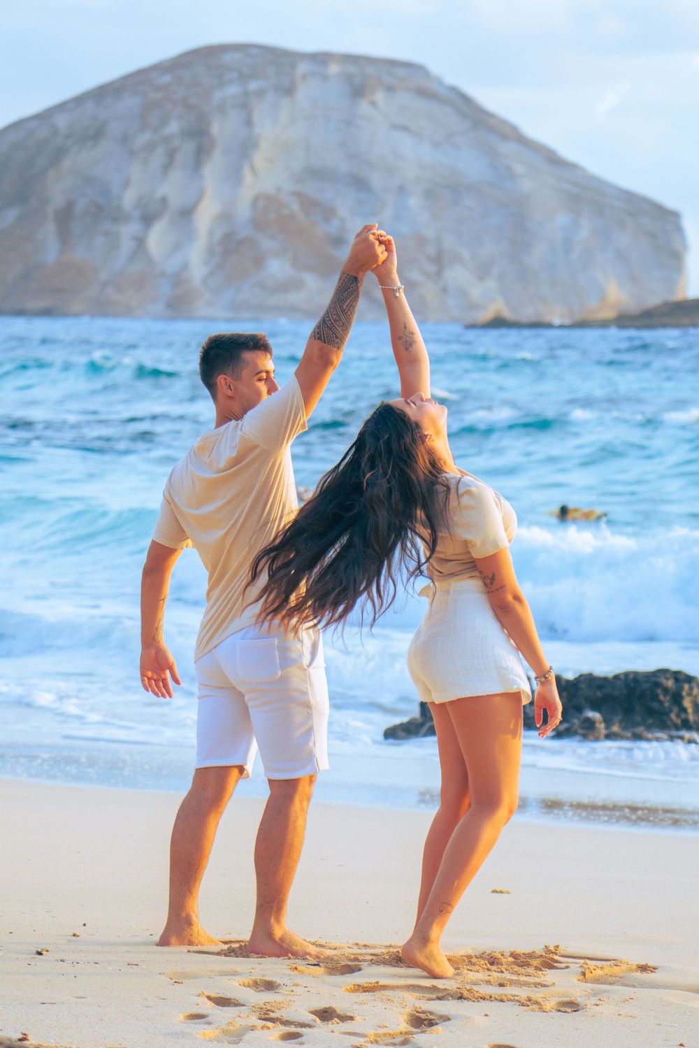 Couple dancing together on a sunny beach with turquoise waves and rocky backdrop.