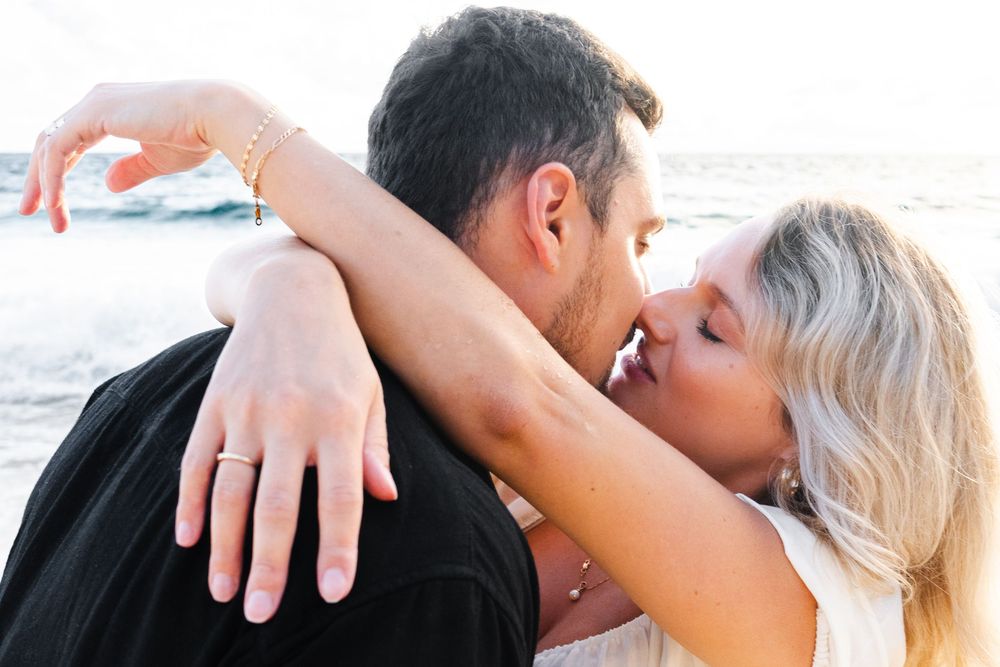 Couple sharing passionate kiss on beach, woman showing engagement ring while embracing partner.