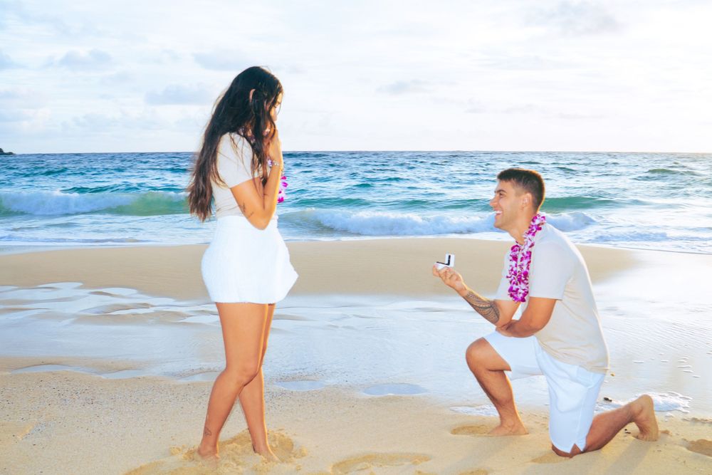 Man kneeling on a beach proposing to woman with engagement ring at sunset.