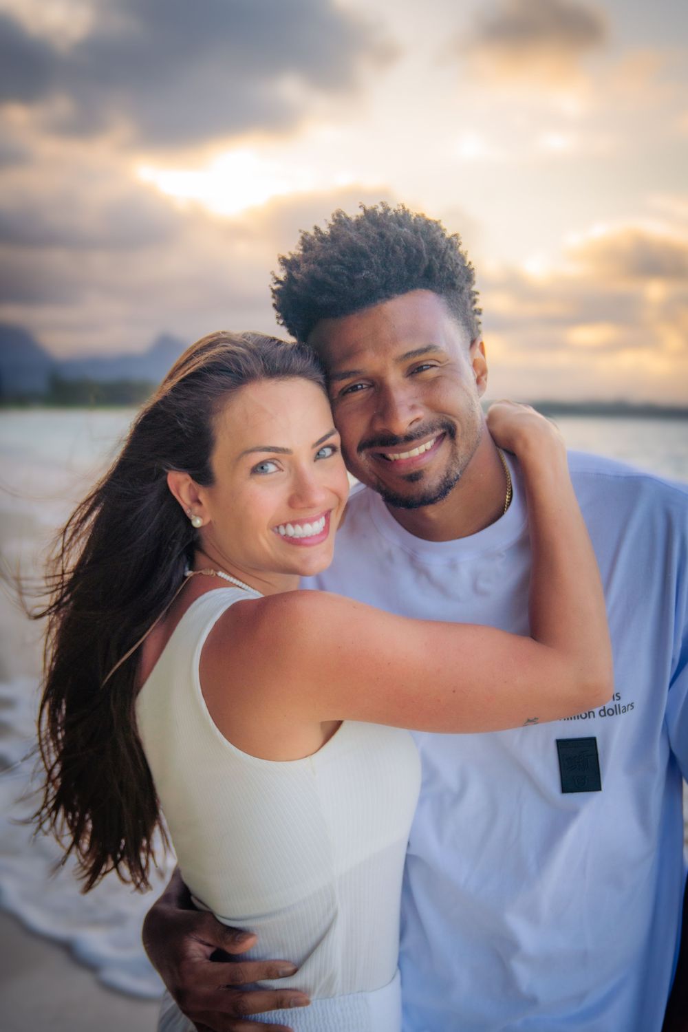 Happy couple embracing on a beach at sunset, smiling warmly with ocean and dramatic sky in the background.