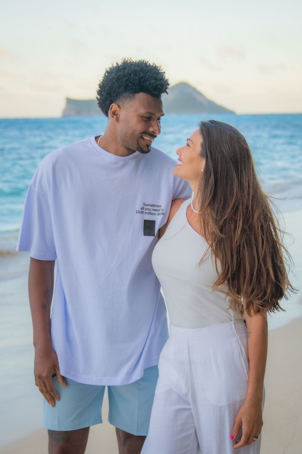 A couple smiles at each other on a tropical beach, man in white tee and shorts, woman in white outfit, ocean backdrop.