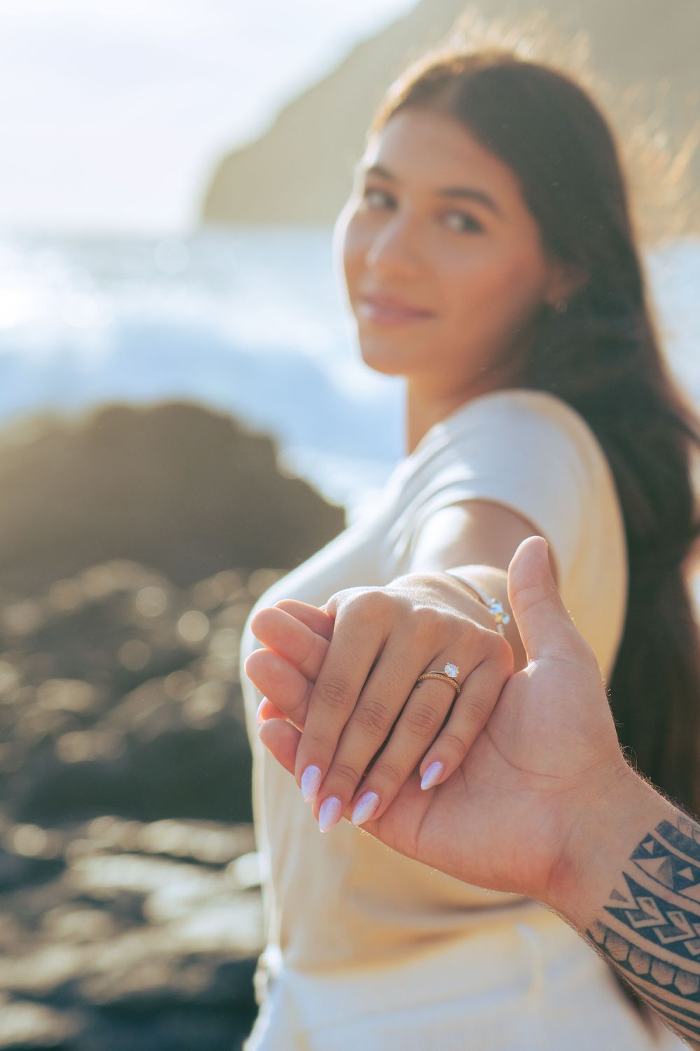 Woman showing engagement ring on hand while holding partners tattooed hand at beach.