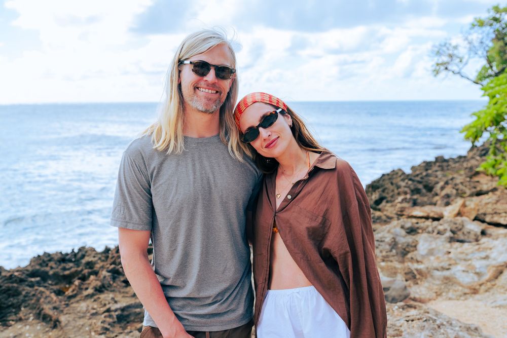 Smiling couple wearing sunglasses posing together on a rocky Hawaiian coastal overlook.