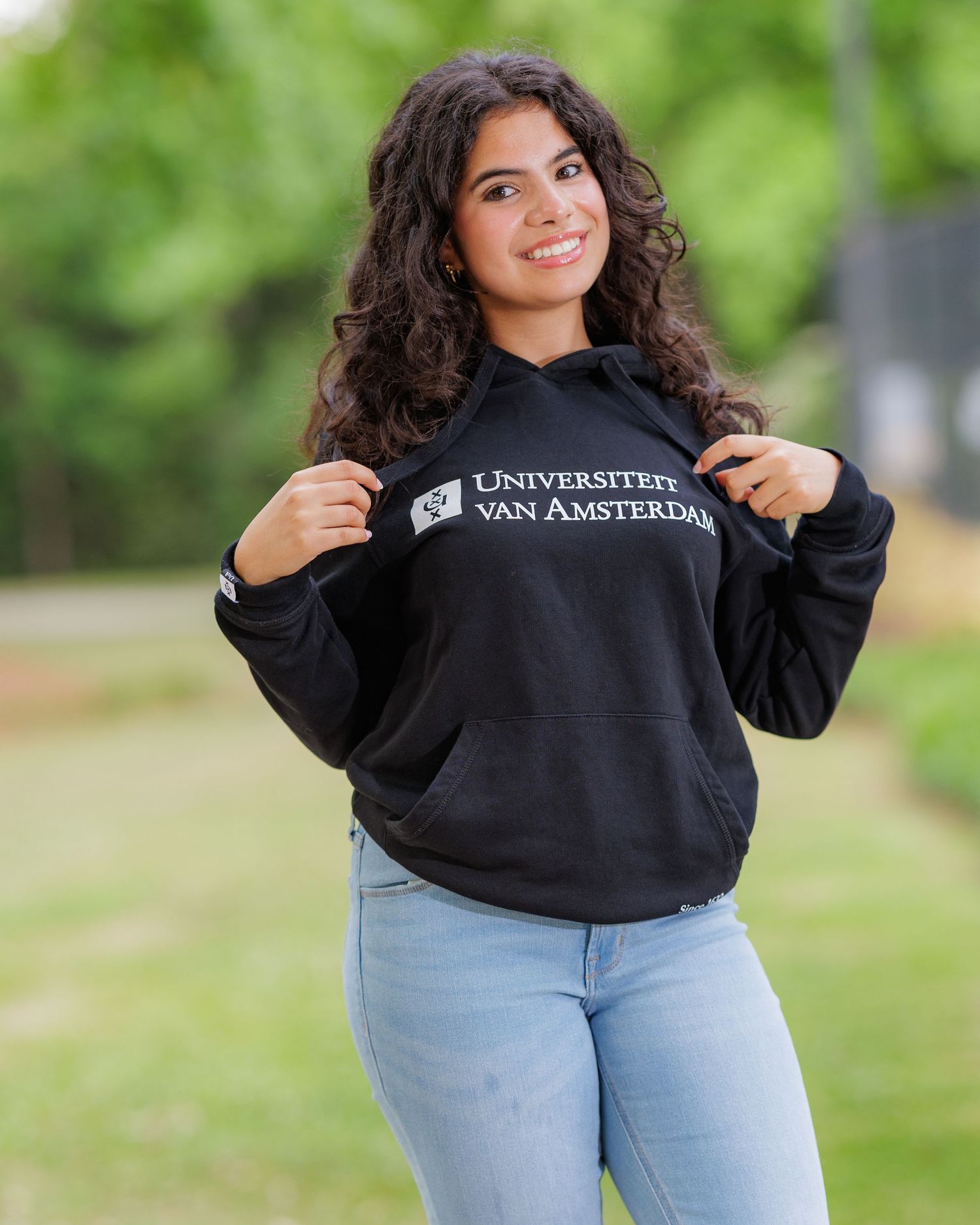 Young woman smiling outdoors wearing a black University of Amsterdam hoodie and light blue jeans.