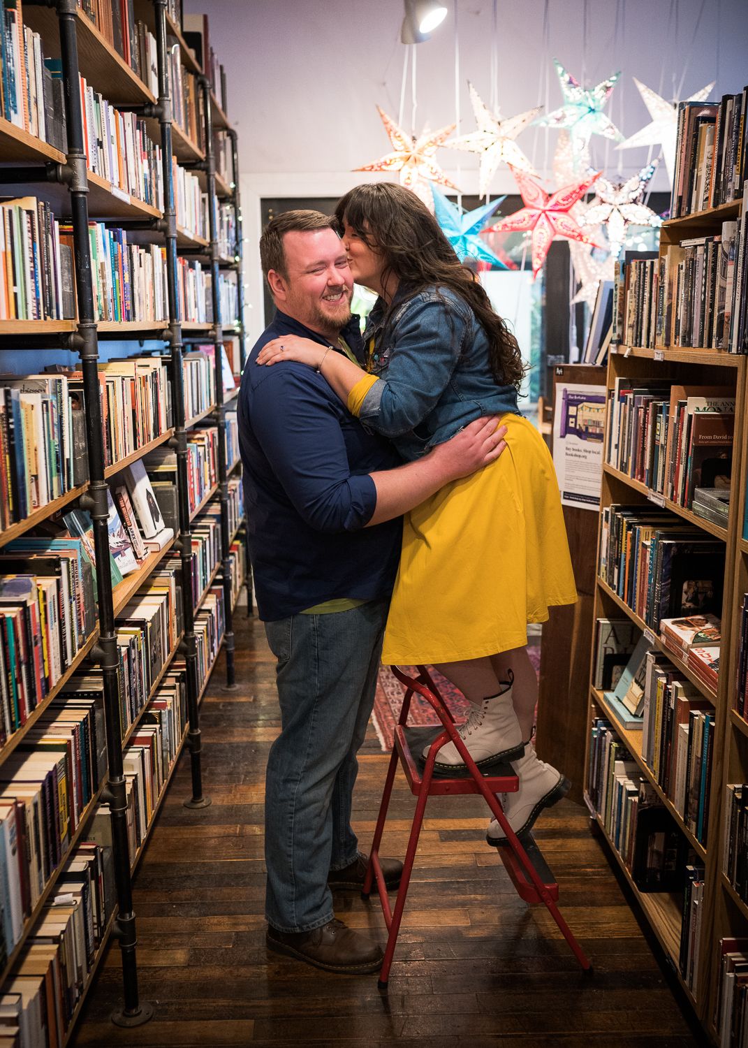 Couple embracing romantically in a cozy bookstore aisle, woman in yellow dress standing on red ladder, colorful star decorations above.