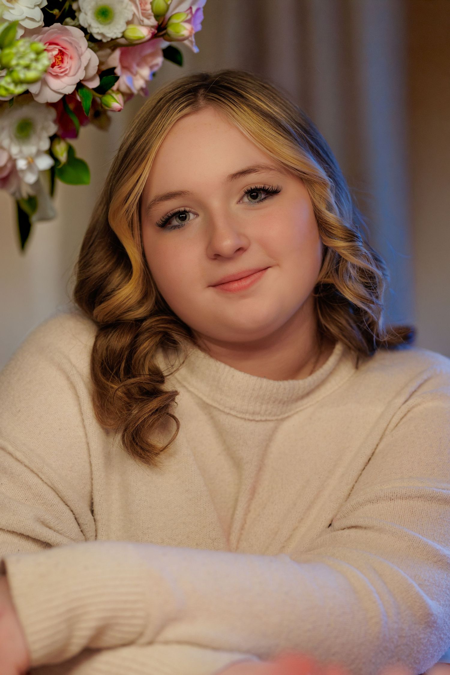 Young blonde woman in cream sweater smiling warmly, seated near a floral arrangement with pink and white flowers.