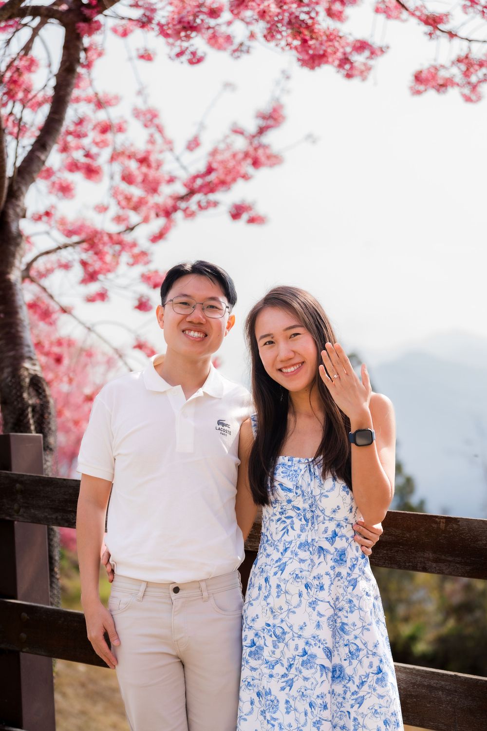 Happy couple posing under pink cherry blossom tree, woman showing engagement ring, wearing blue floral dress outdoors.