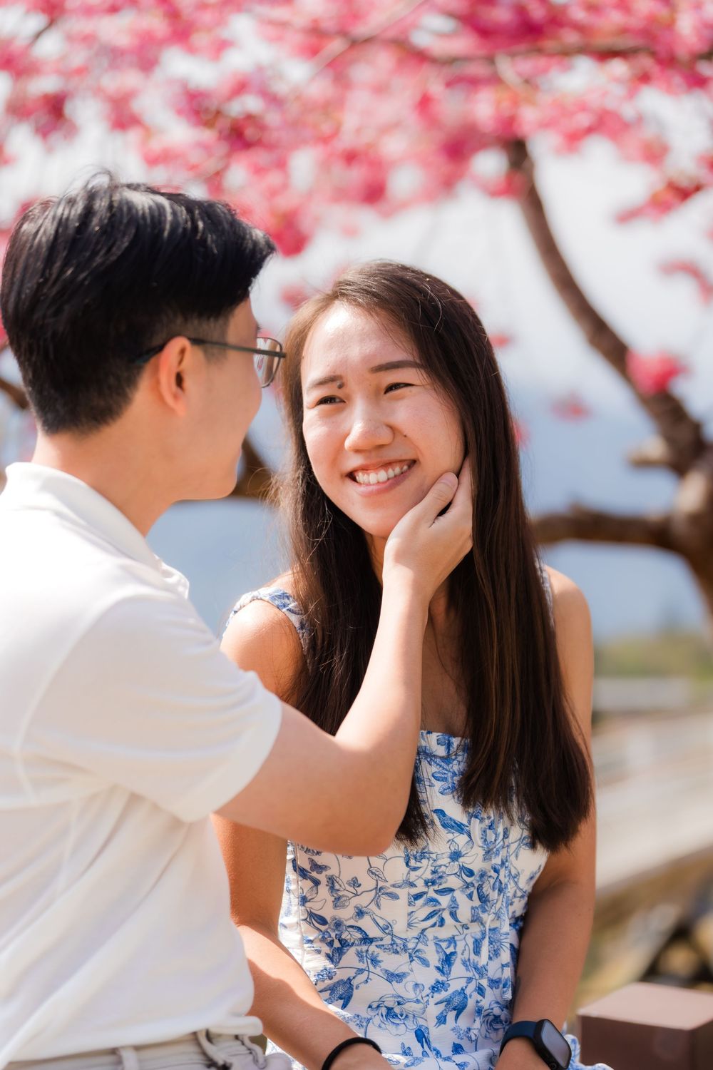 A smiling Asian couple shares a tender moment under pink cherry blossom trees, man gently touching woman's face.