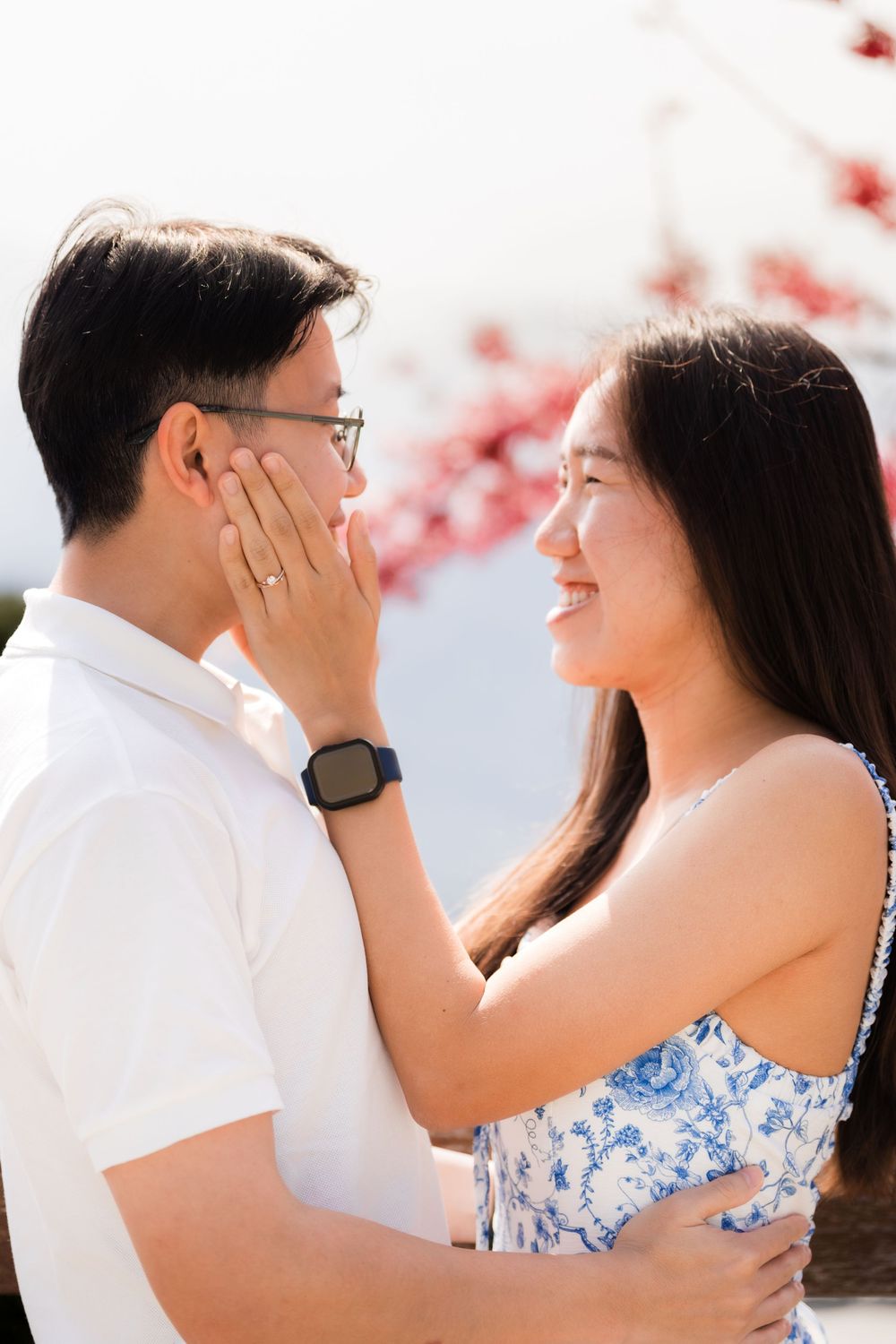 A smiling Asian couple shares a tender moment outdoors, with the woman gently touching the man's face near pink flowers.