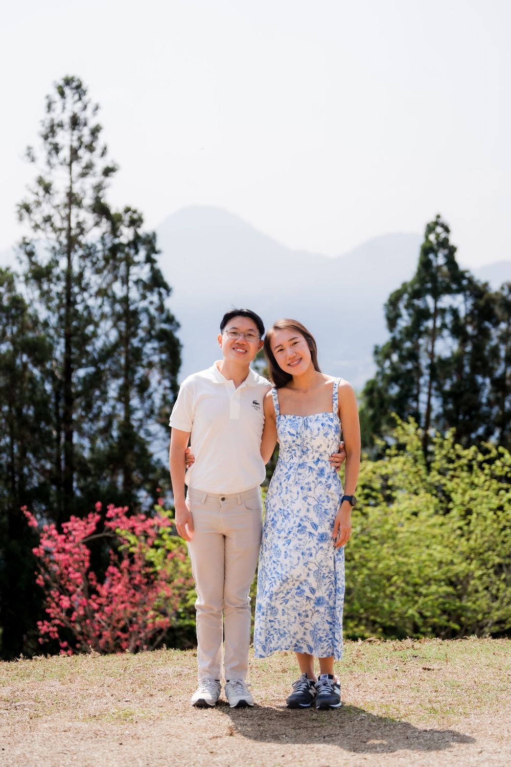 Asian couple smiling together outdoors with tall trees and misty mountains in the background.