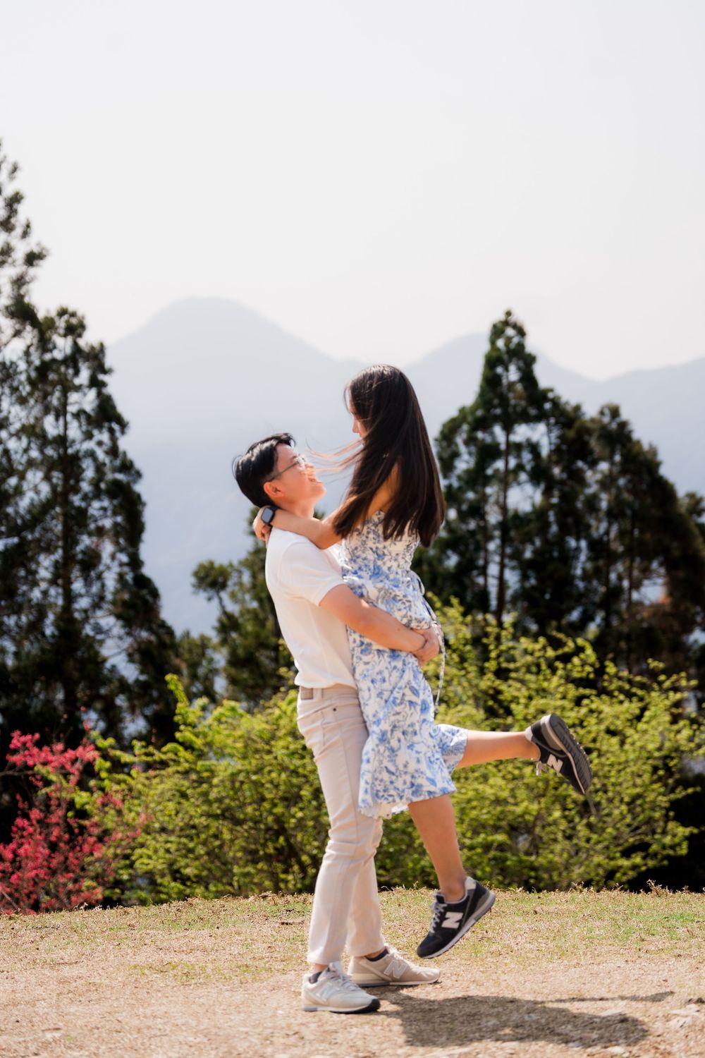 Man lifting woman in his arms outdoors with lush green trees and mountain scenery behind them.