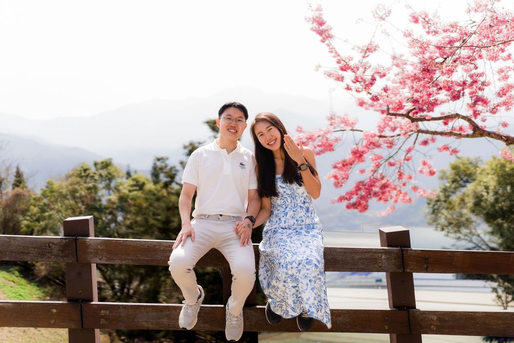 Couple sitting on a wooden fence under a pink cherry blossom tree with mountains in the background.