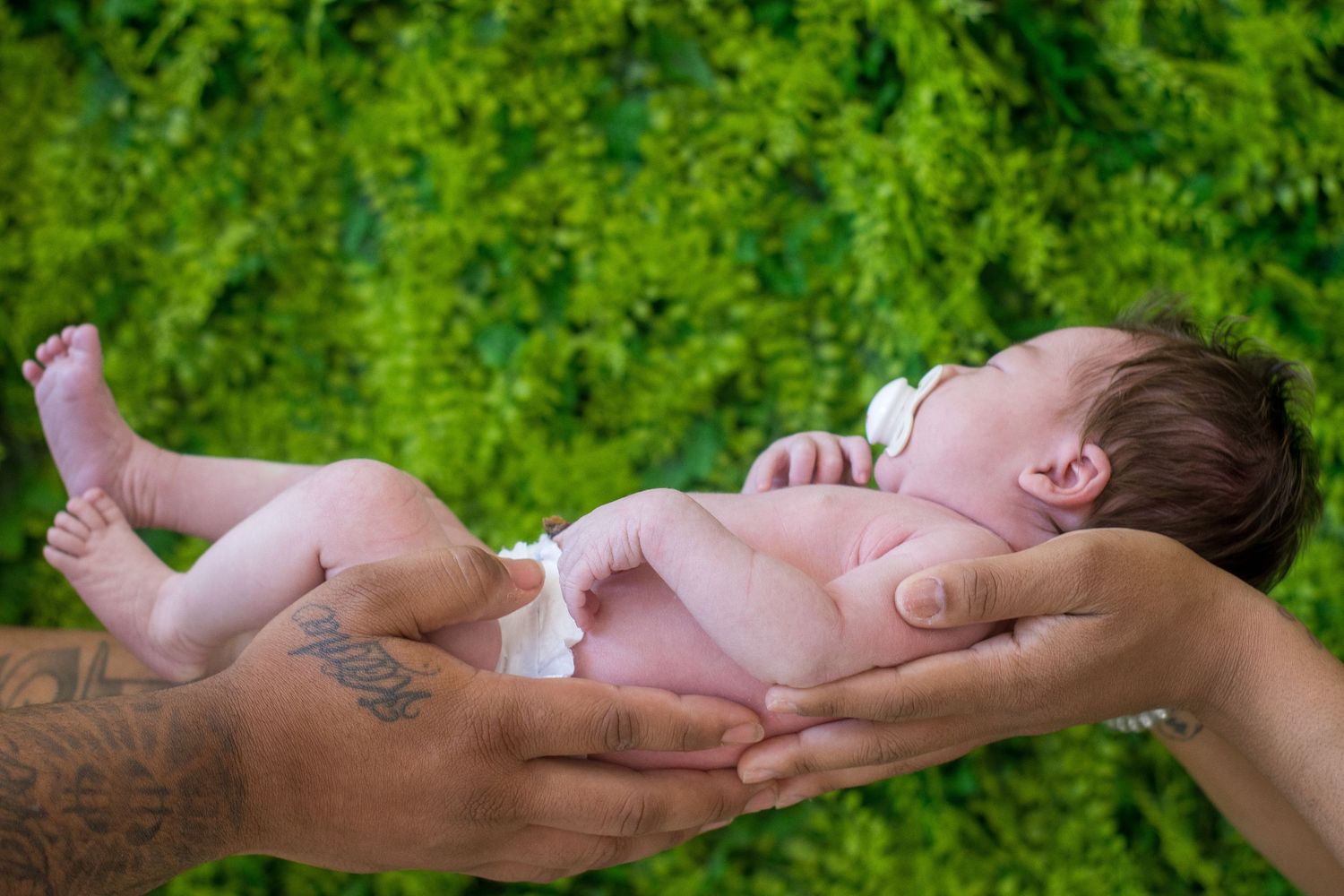 Newborn baby sleeping peacefully in tattooed parents hands against lush green background.