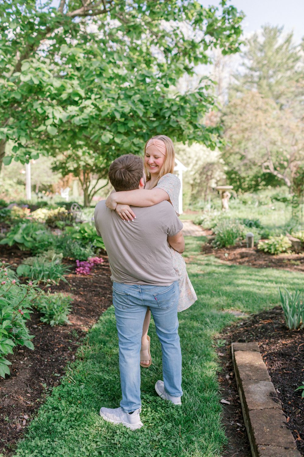A couple embraces in a lush garden path, the woman smiling down at the man lifting her among green plants.