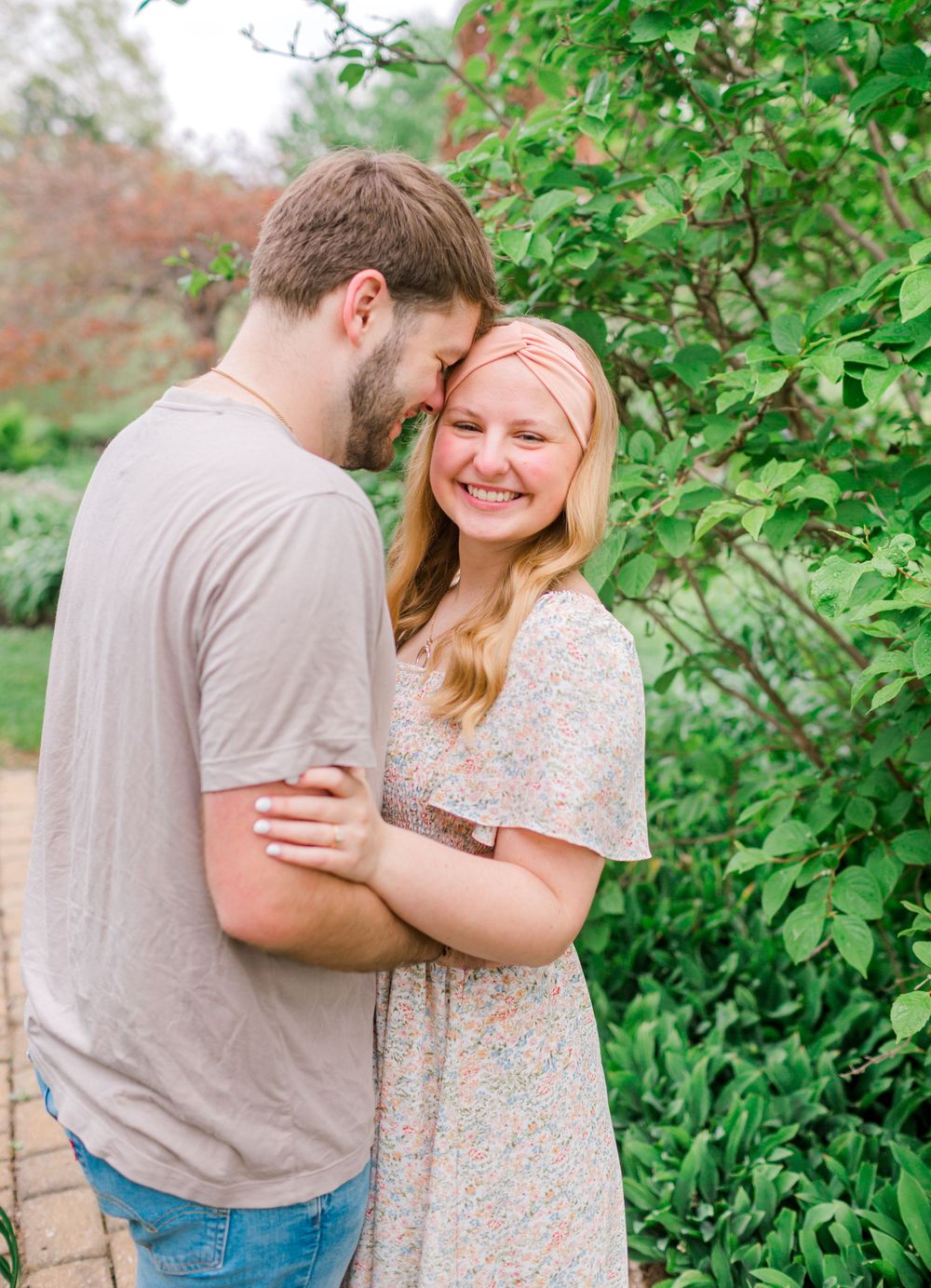 A couple shares a tender moment outdoors, smiling together near lush green bushes, wearing casual spring attire.