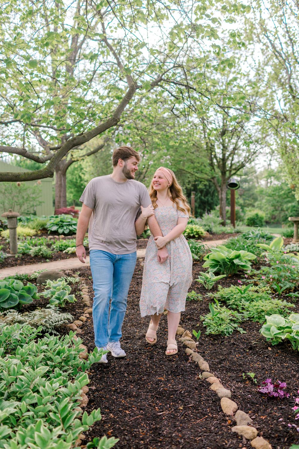 A couple walks hand-in-hand through a lush garden path, smiling at each other among green plants and blooming trees.