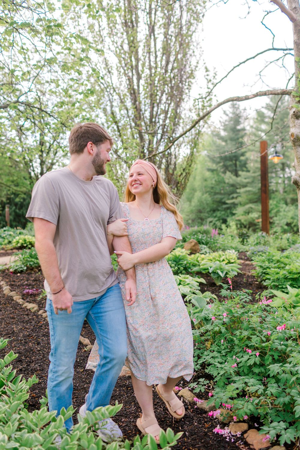 A smiling couple walks through a lush garden, the woman in a floral dress and the man in a gray tee and jeans.