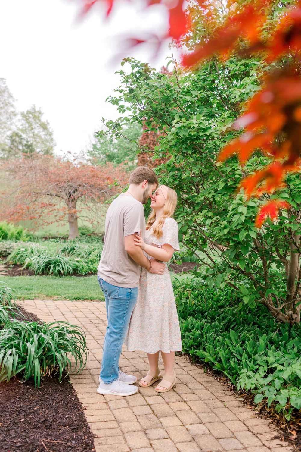 Couple sharing a romantic kiss on a brick garden path surrounded by lush green and red foliage.