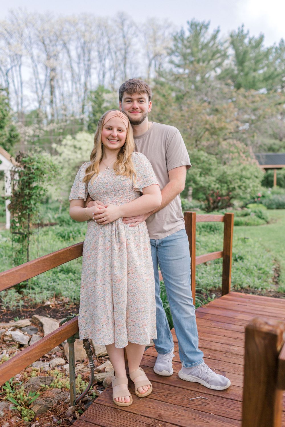 A smiling couple poses on a wooden deck surrounded by lush greenery, the man embracing the woman from behind.