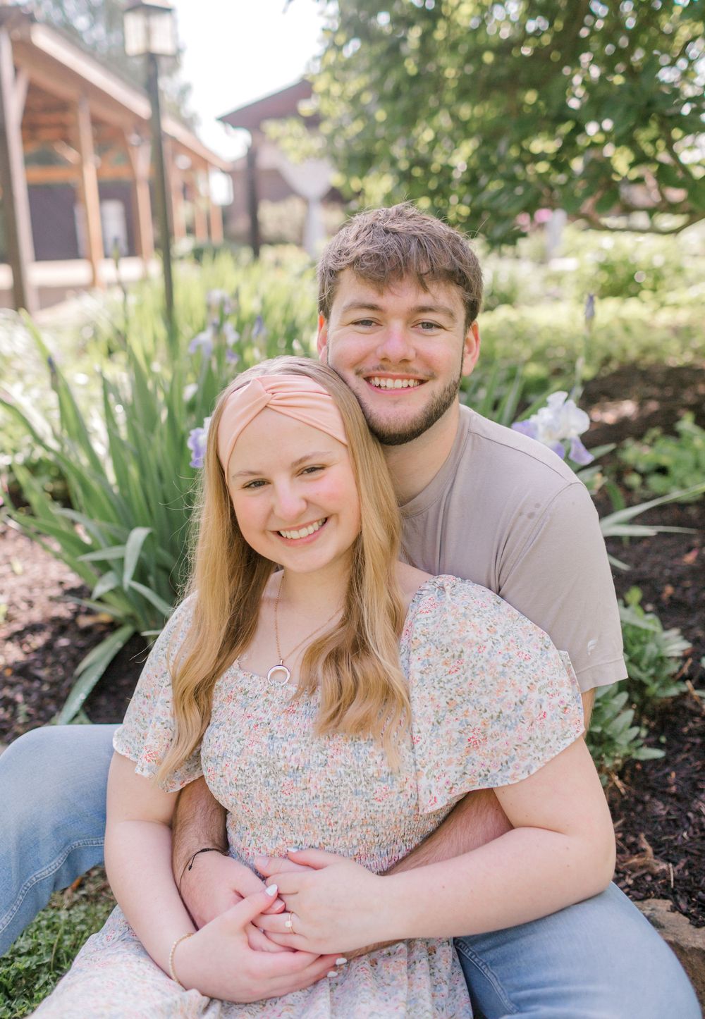A smiling young couple poses together in a lush garden, woman in floral dress, man in tan shirt embracing her.