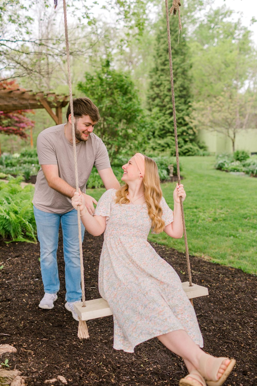 A couple shares a joyful moment outdoors, woman sitting on a rope swing in a floral dress while man pushes her in a lush garden in Indiana.
