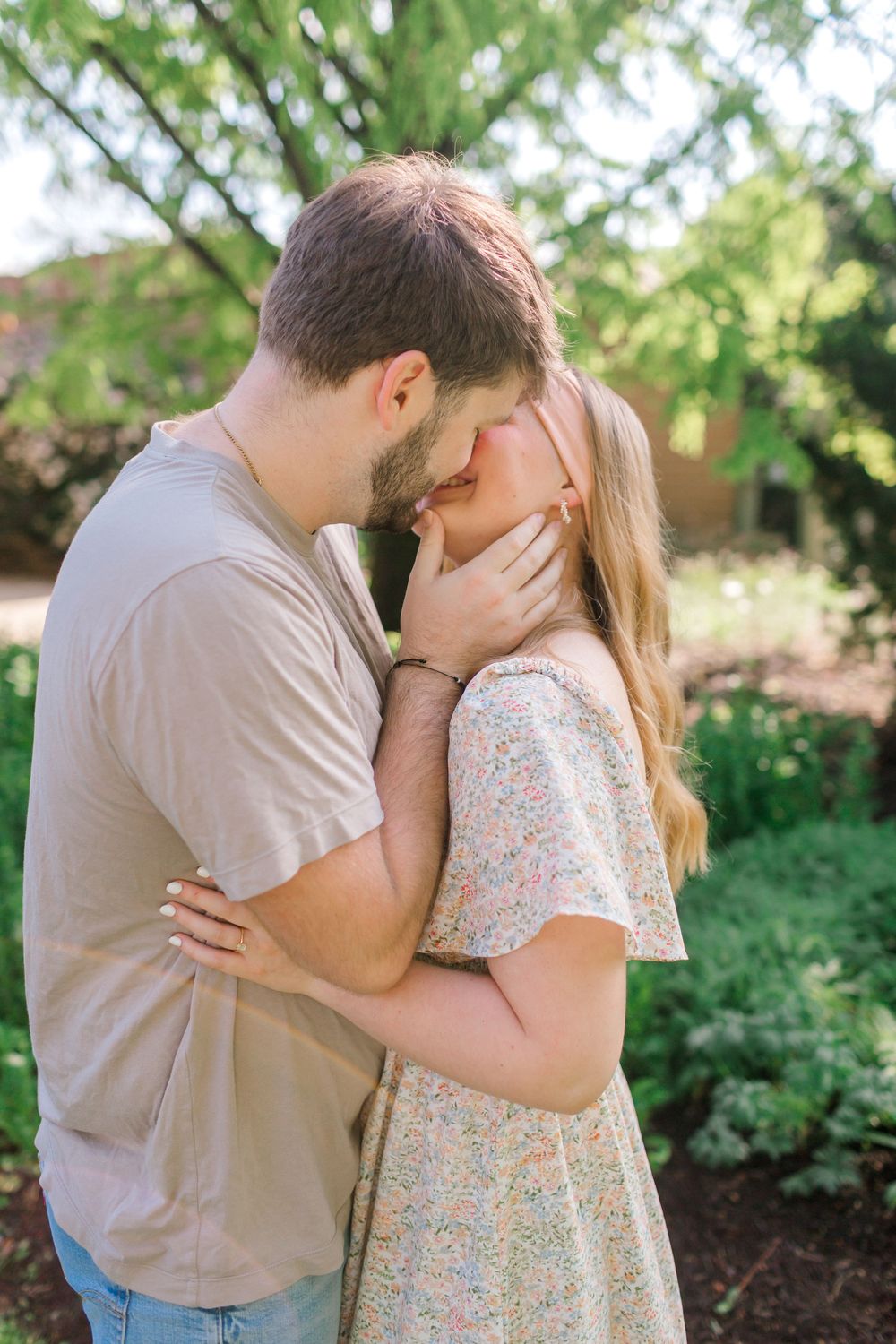 A couple shares a tender kiss outdoors, surrounded by lush green trees and gardens on a sunny day in Indiana.