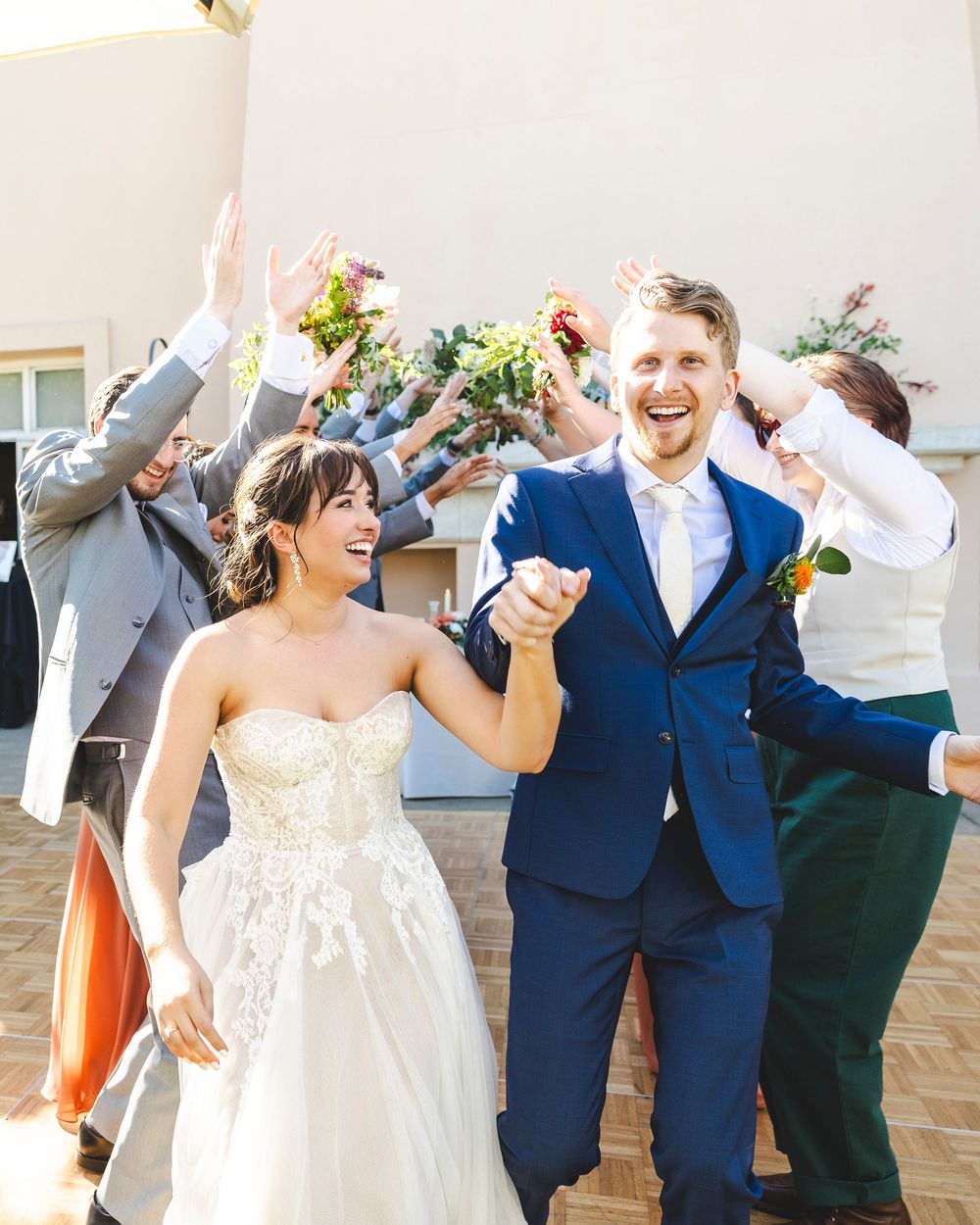 Joyful newlyweds walk through celebratory arch of raised hands with wedding party outdoors.