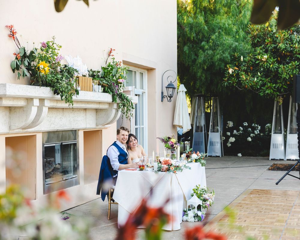 Bride and groom seated at elegant outdoor sweetheart table adorned with floral centerpieces at venue.