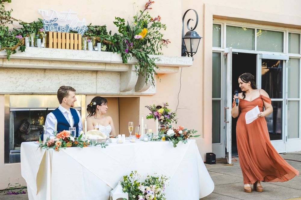 Bridesmaid gives speech at outdoor wedding reception while couple sits at floral sweetheart table.