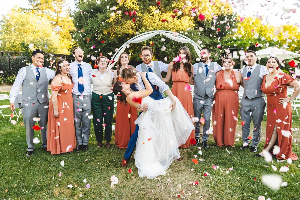 A wedding party celebrates with confetti toss as groom dips bride under a floral arch outdoors.