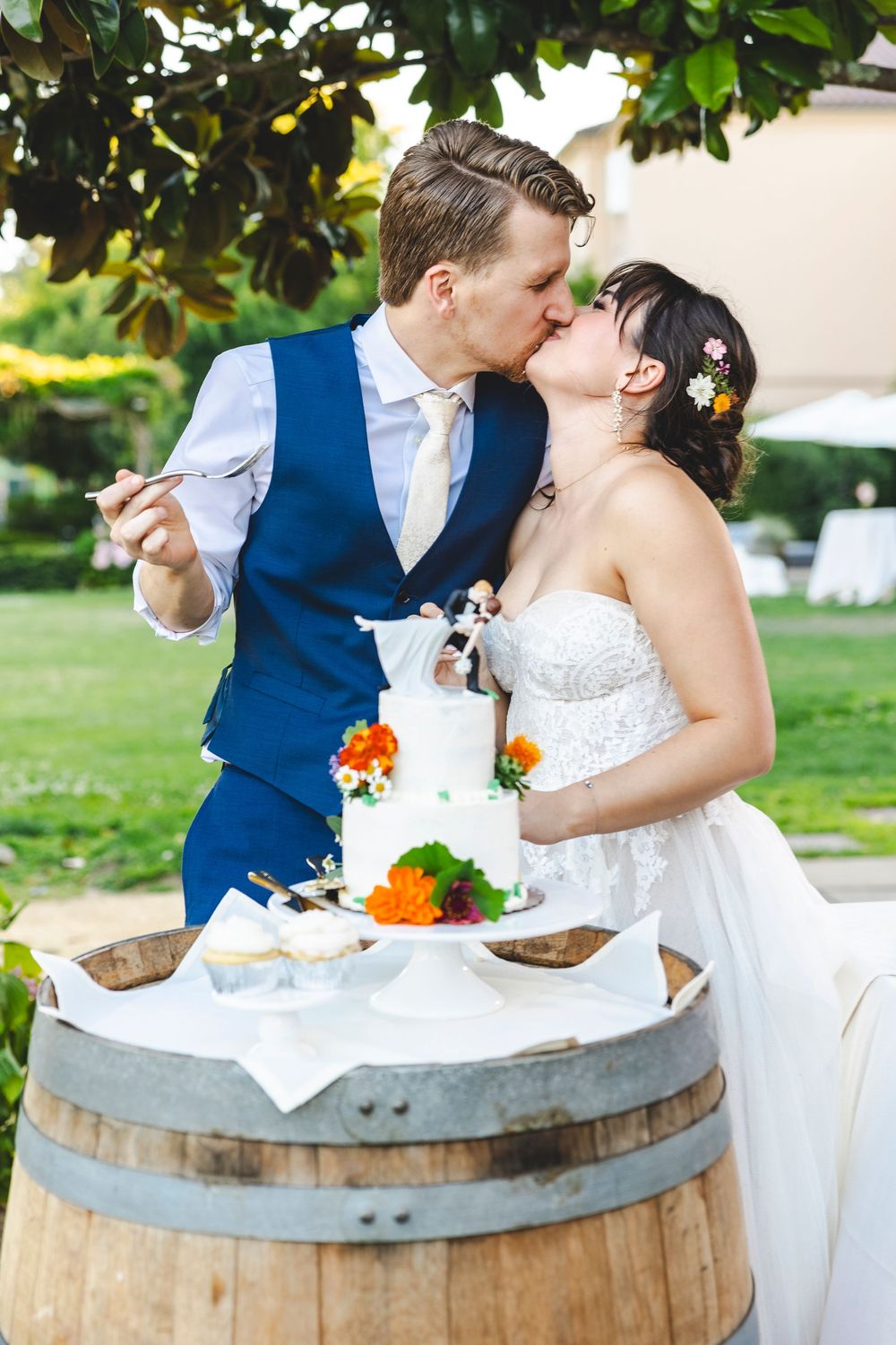 Bride and groom kissing while cutting their white wedding cake decorated with orange flowers on a wooden barrel outdoors.